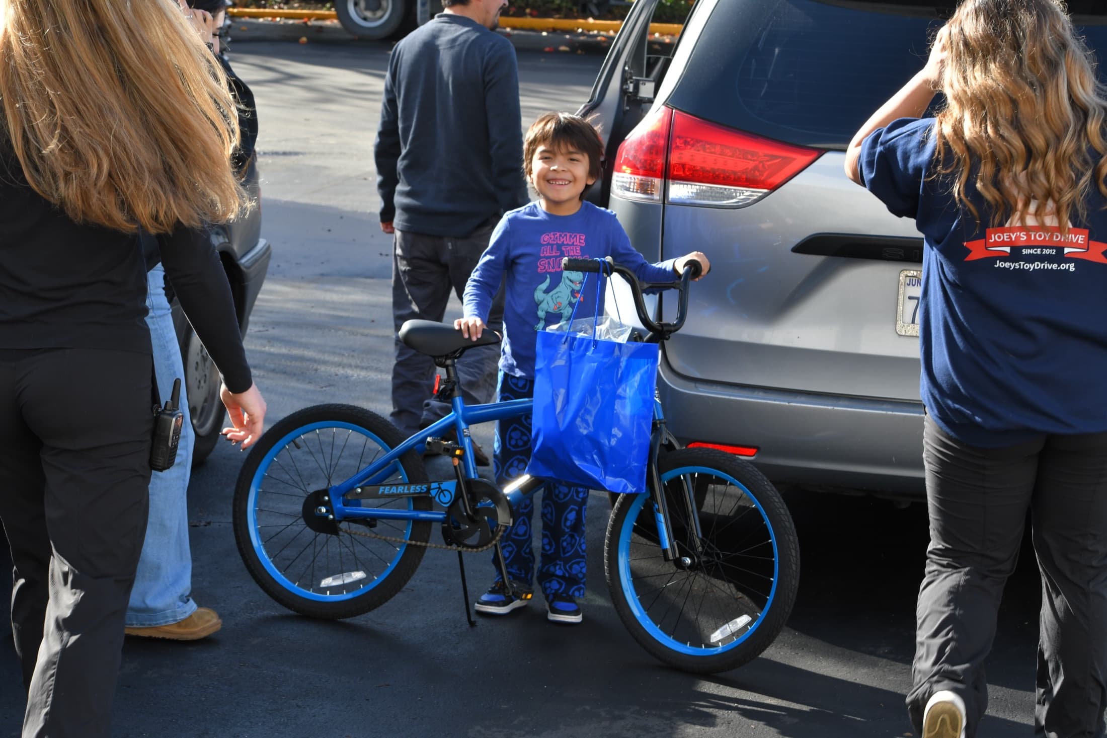 A smiling boy holds the handlebars of his new blue bike, surrounded by Joey's Toy Drive volunteers on distribution day
