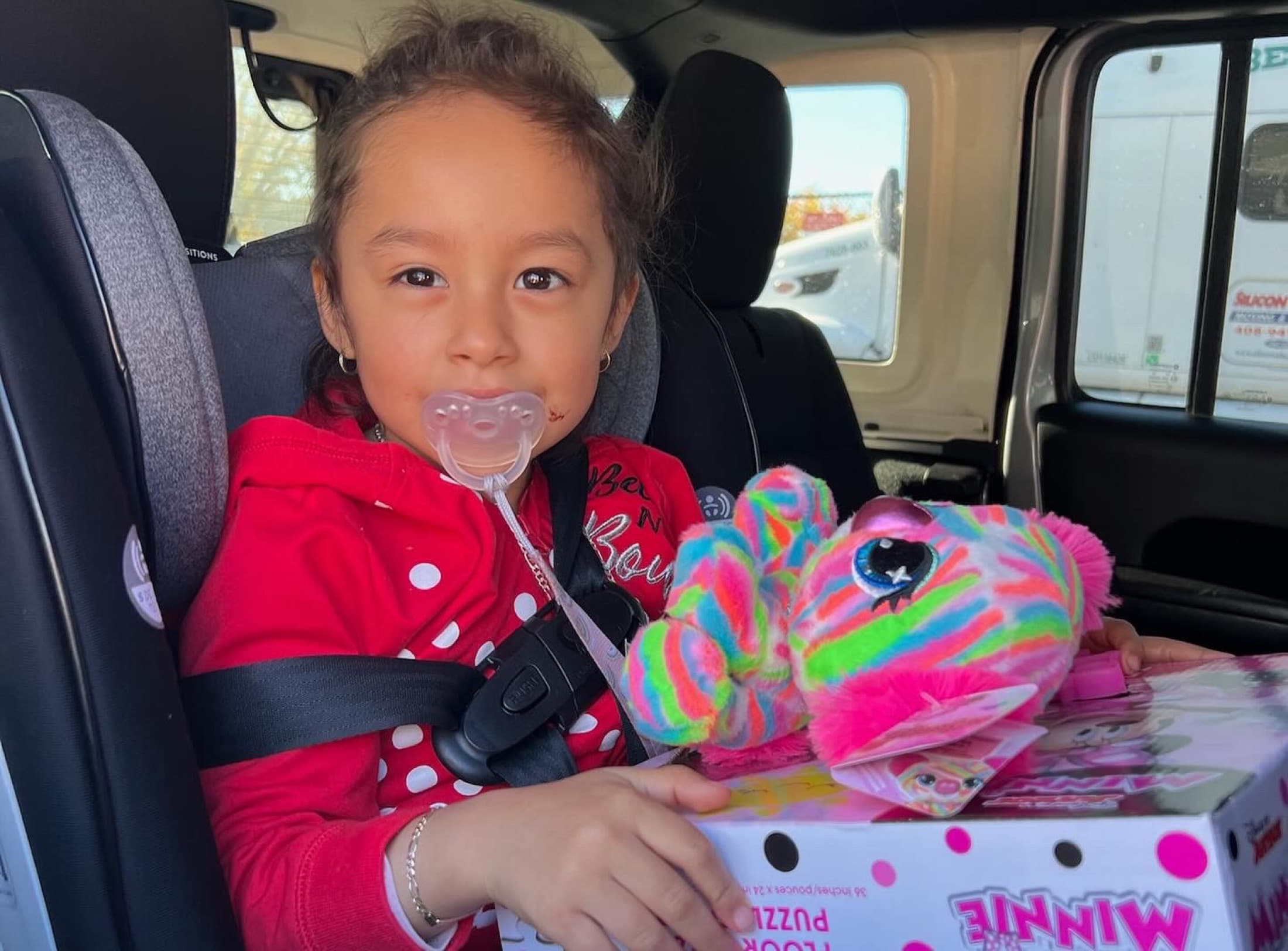A little girl buckled into her car seat, hugging a bright striped stuffed animal on top of a puzzle box