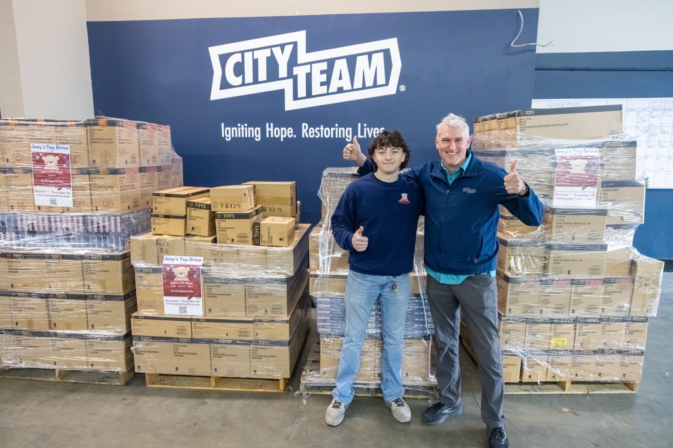 Two people give a thumbs-up in front of pallets of boxed toys inside the CityTeam warehouse
