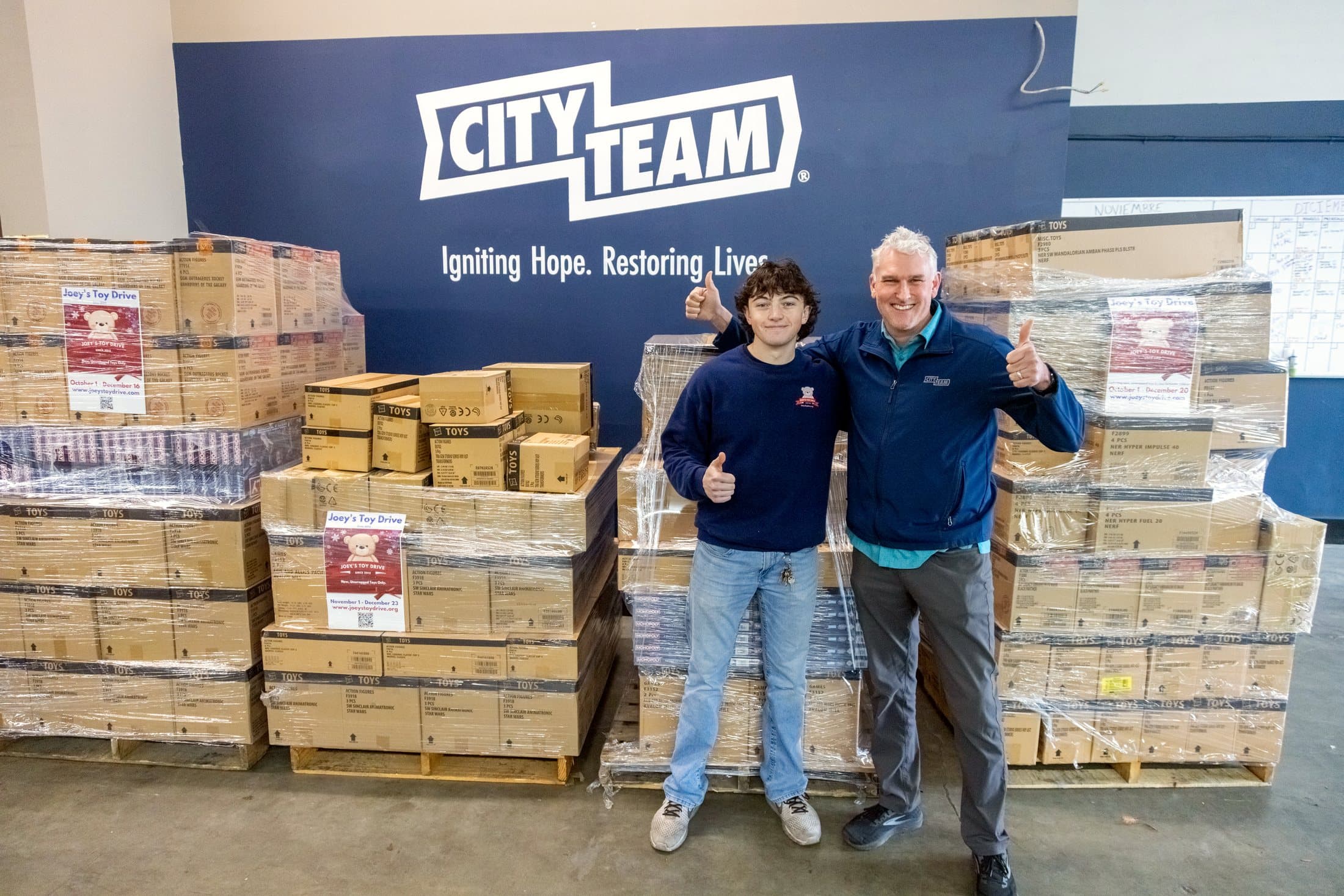 Volunteers standing beside pallets of boxed toys labeled for Joey's Toy Drive at a Bay Area distribution partner