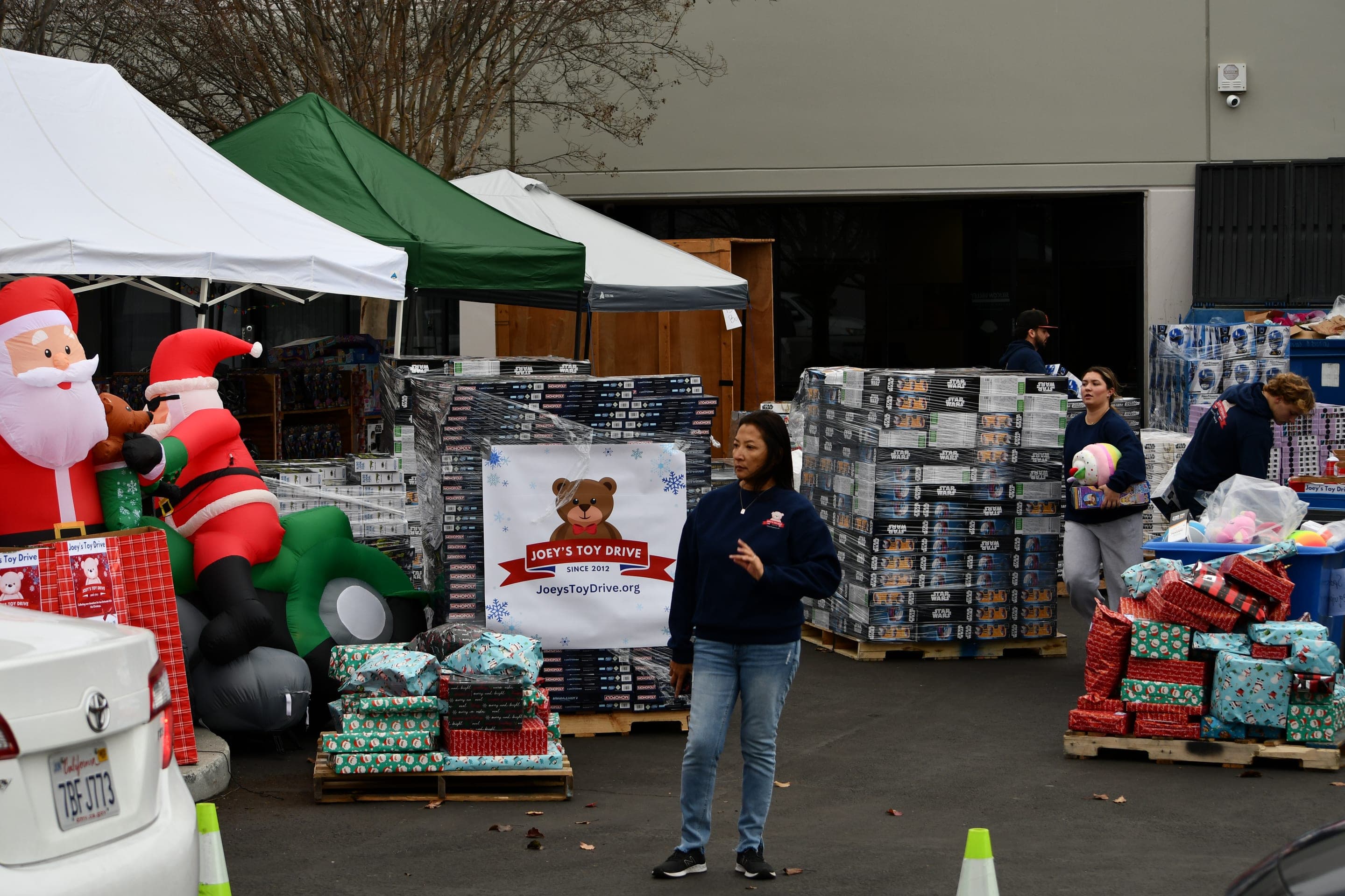Volunteers in Joey's Toy Drive sweatshirts working among shrink-wrapped pallets of boxed toys and stacks of wrapped gifts outside the San Jose warehouse