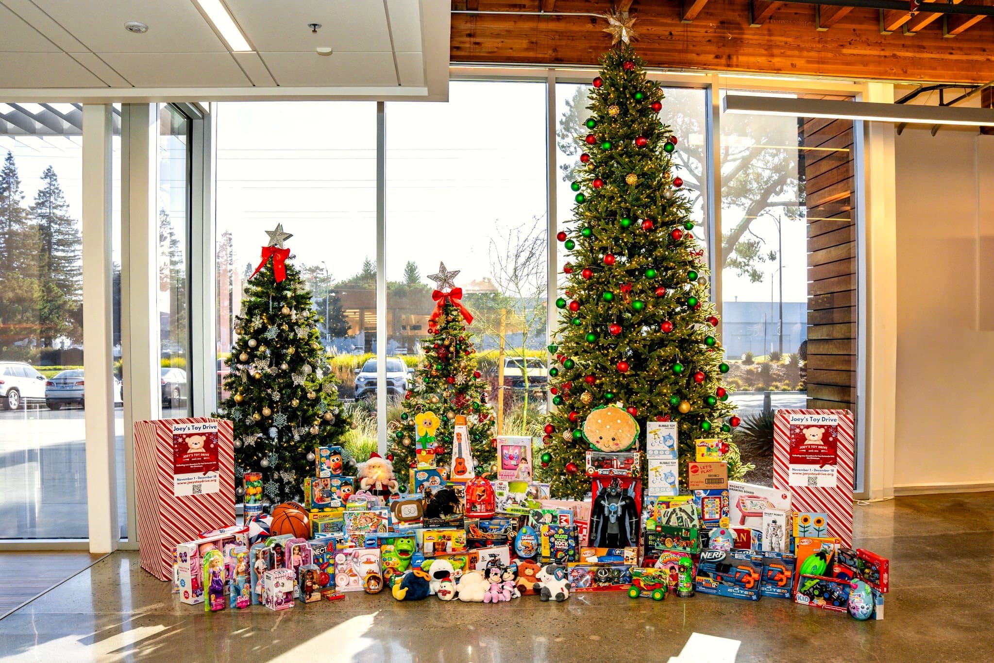 Donated toys spread across an office lobby floor beneath three decorated Christmas trees