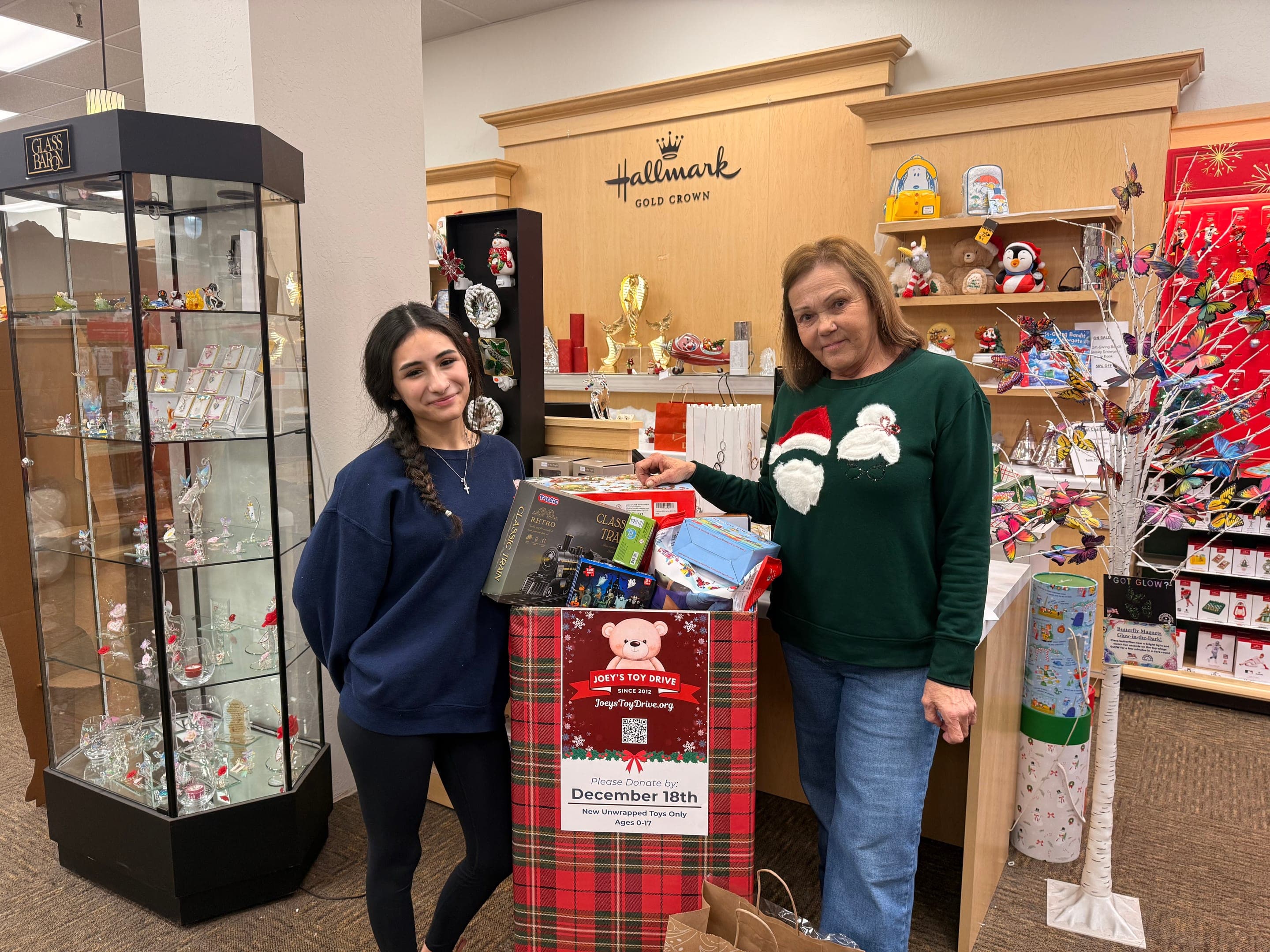 A Joey's Toy Drive collection box filled with toys inside a gift shop, with two people standing beside it