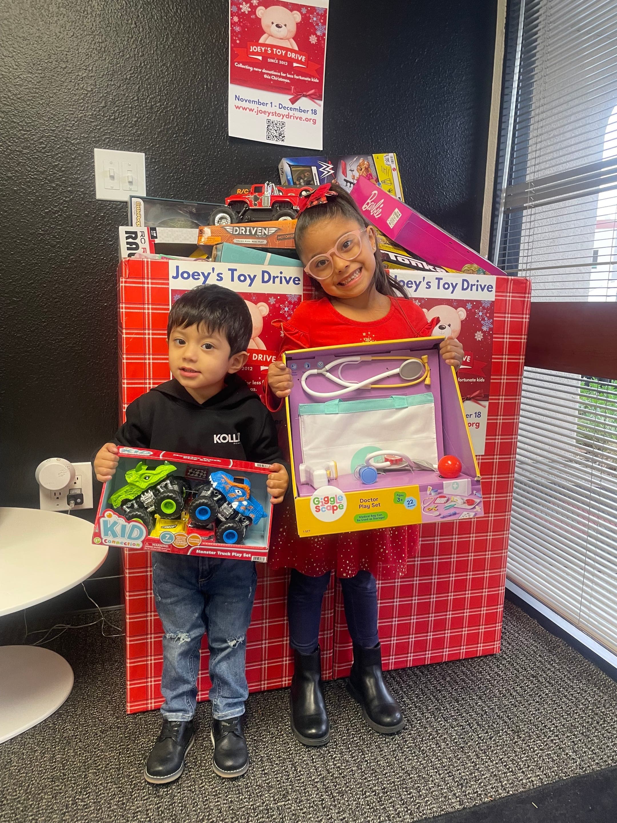 Two children holding the new toys they just received next to a Joey's Toy Drive collection box