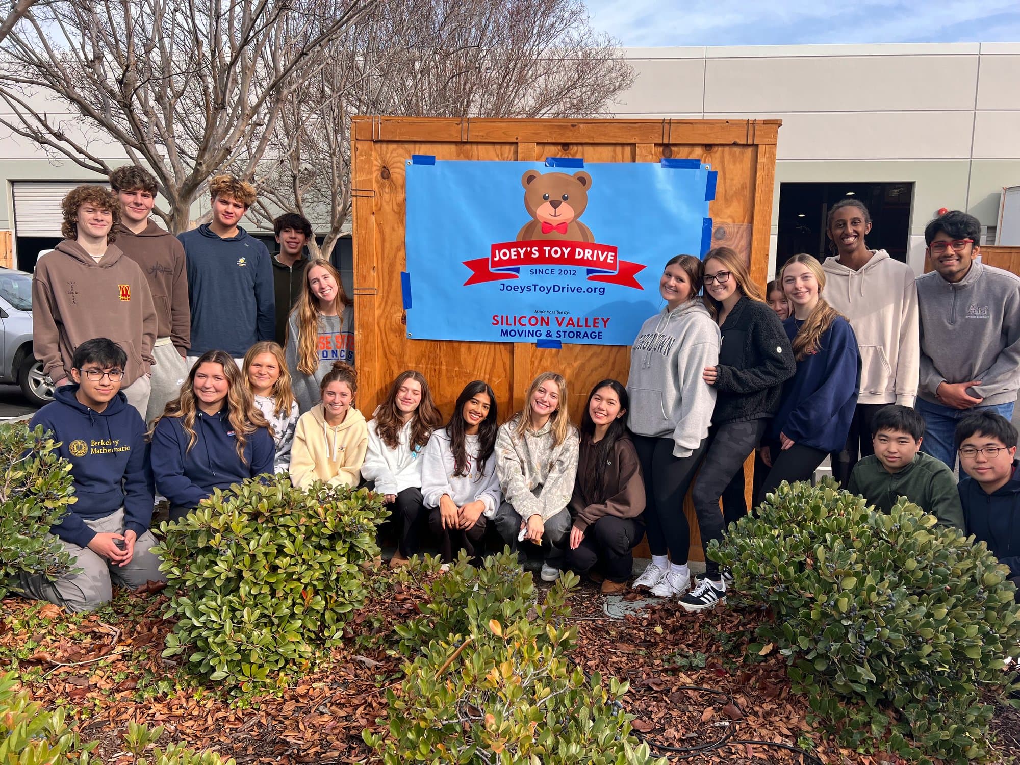 A large group of student volunteers gathered in front of the Joey's Toy Drive and Silicon Valley Moving banner outside the warehouse