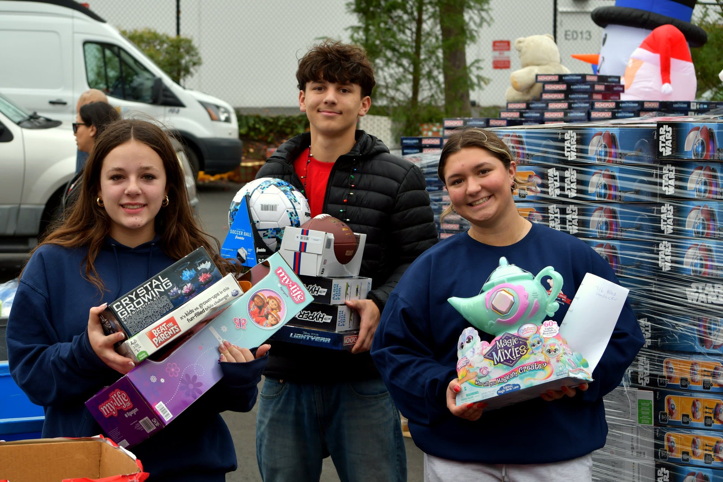 Three young volunteers holding new toys on a Joey's Toy Drive collection day, with pallets of donated gifts stacked behind them