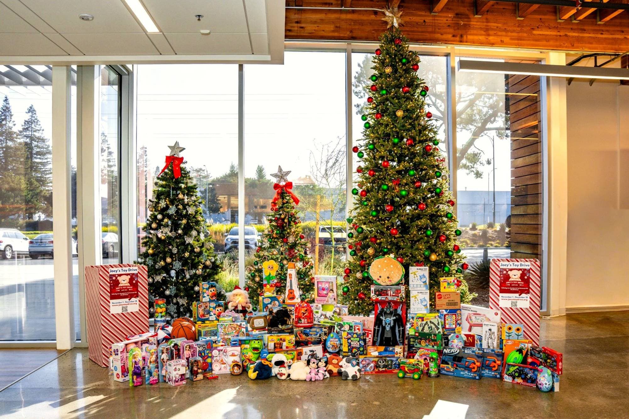 Two Joey's Toy Drive collection boxes flanking three decorated Christmas trees and a wide spread of donated toys in an office lobby