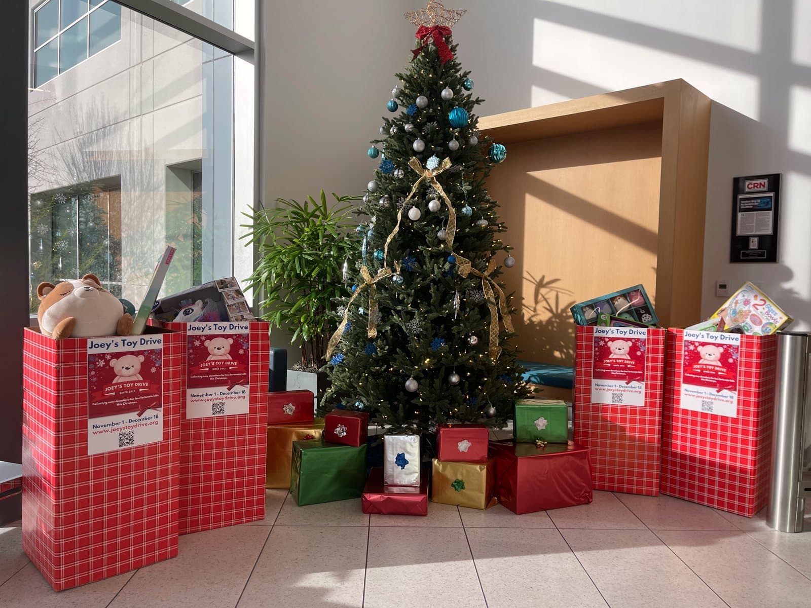 Four Joey's Toy Drive collection boxes beside a decorated tree in an office lobby, wrapped gifts already stacked underneath