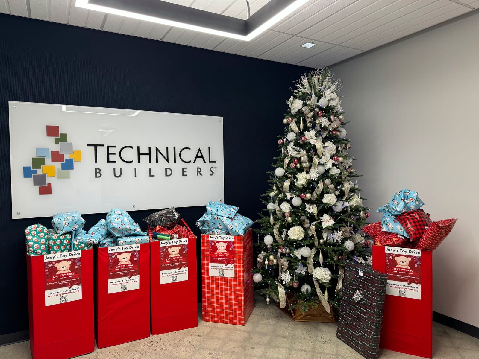 Joey's Toy Drive collection boxes filling with wrapped gifts beside a decorated tree in a corporate lobby